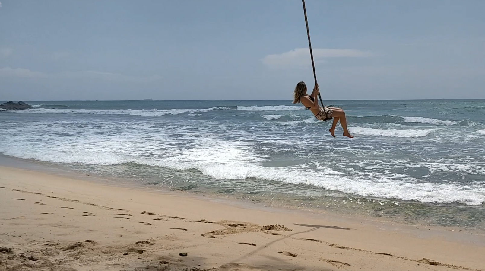 Frau schwingt auf einer Schaukel am Strand hoch Ăźber den Wellen des Meeres.